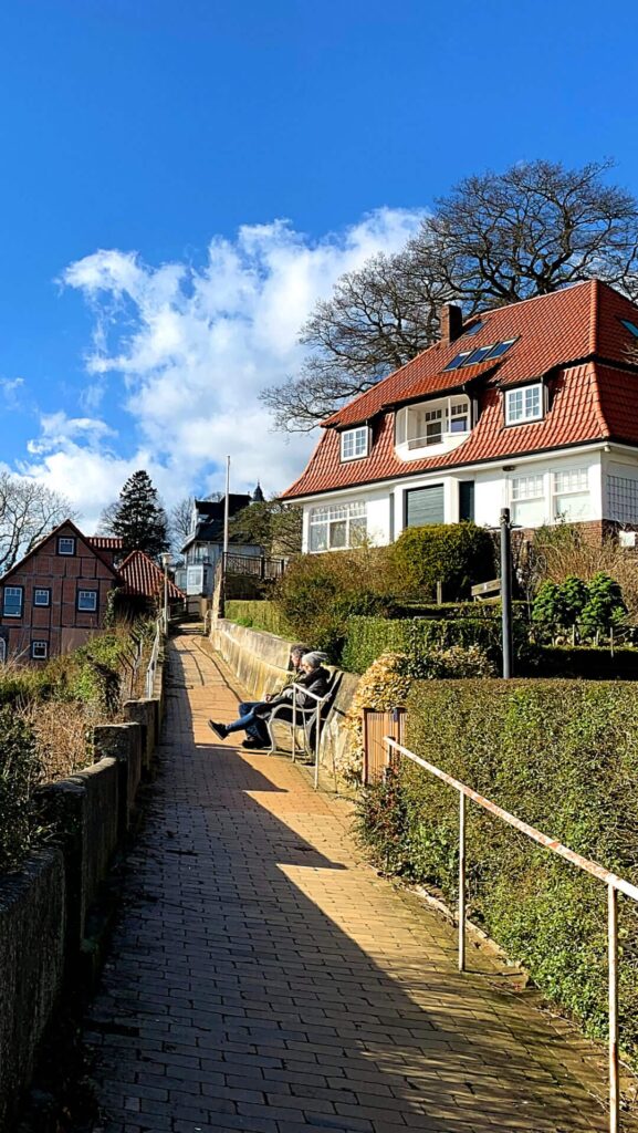 Ein Weg führt zu einer Bank, auf der zwei Menschen sitzen. Dahinter steht ein Haus mit Blick auf die Elbe.