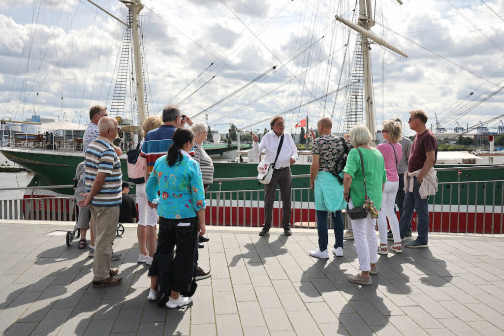 Ein Mann steht in der Mitte einer Gruppe direkt am Hafen vor einem Schiff und erzählt ihnen etwas.