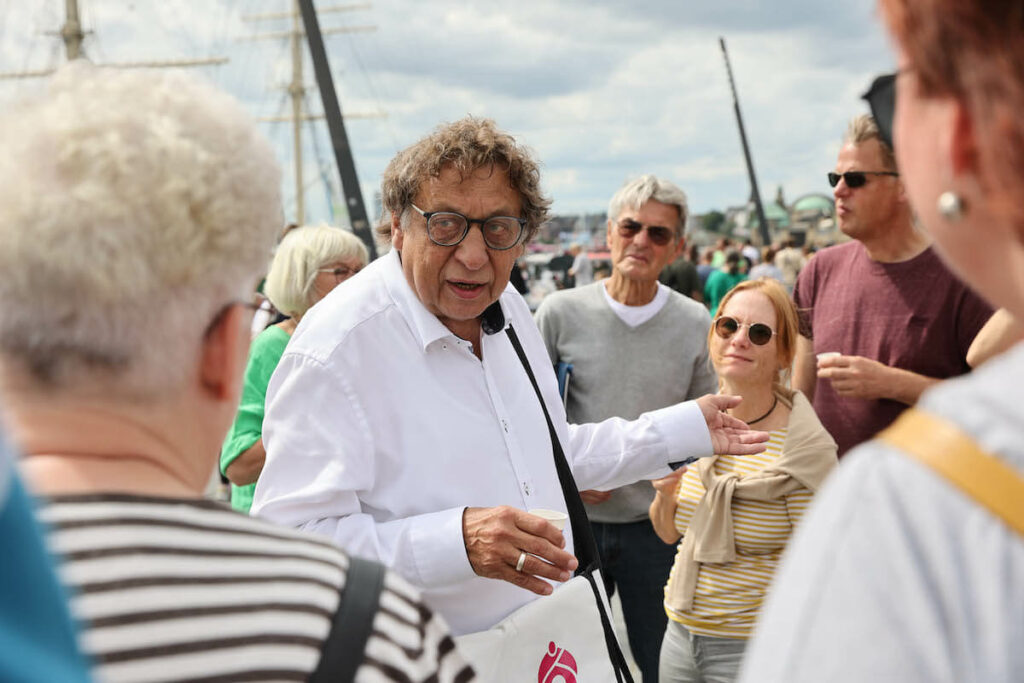 Ein Mann steht in der Mitte einer Gruppe direkt am Hafen und erzählt ihnen etwas.