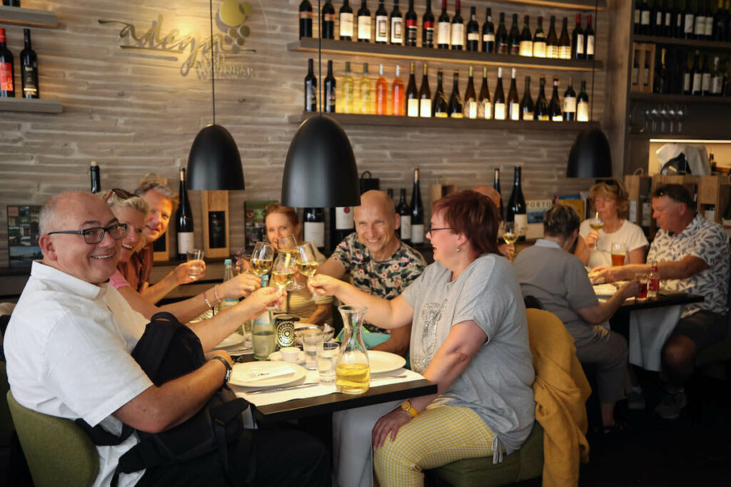 Eine Gruppe von Menschen stößt mit einem Glas Weißwein an und lächelt dabei.