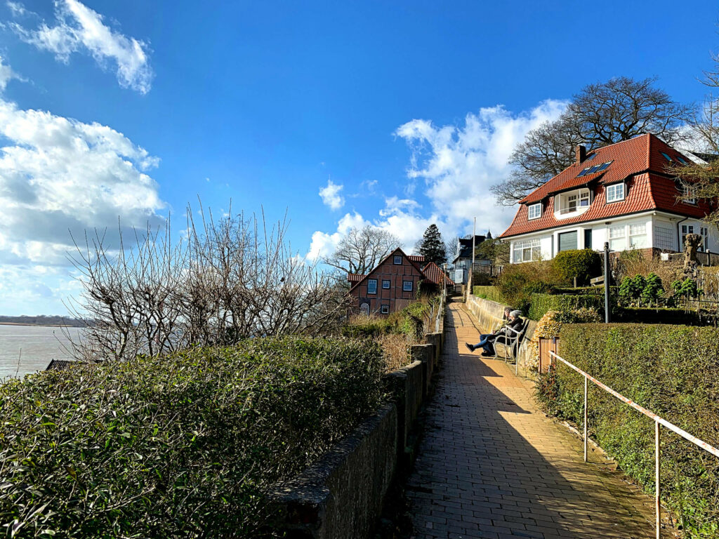 Ein Weg führt zu einer Bank, auf der zwei Menschen sitzen. Dahinter steht ein Haus mit Blick auf die Elbe.